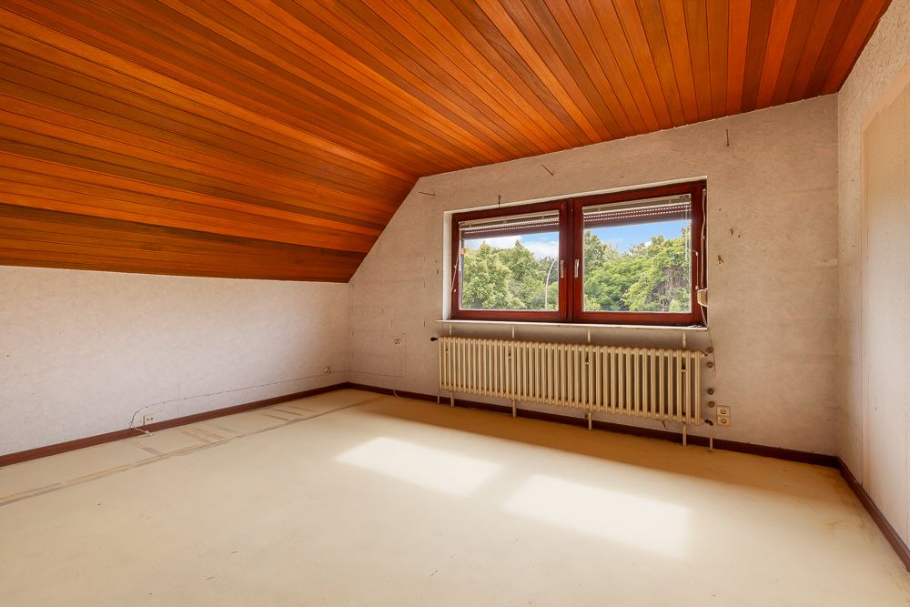 Leeres Zimmer mit schräger Holzdecke, großem Fenster und Heizkörper, Blick auf grüne Bäume.