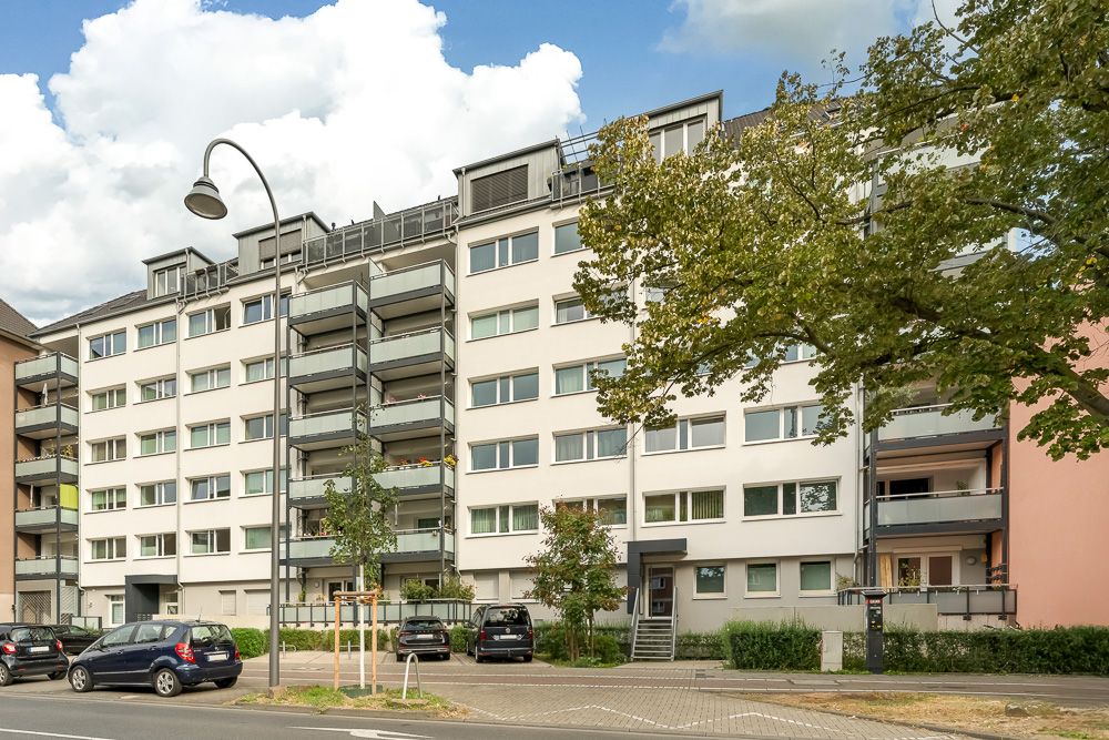 Mehrstöckiges Wohnhaus mit Balkonen, Bäumen und geparkten Autos davor.