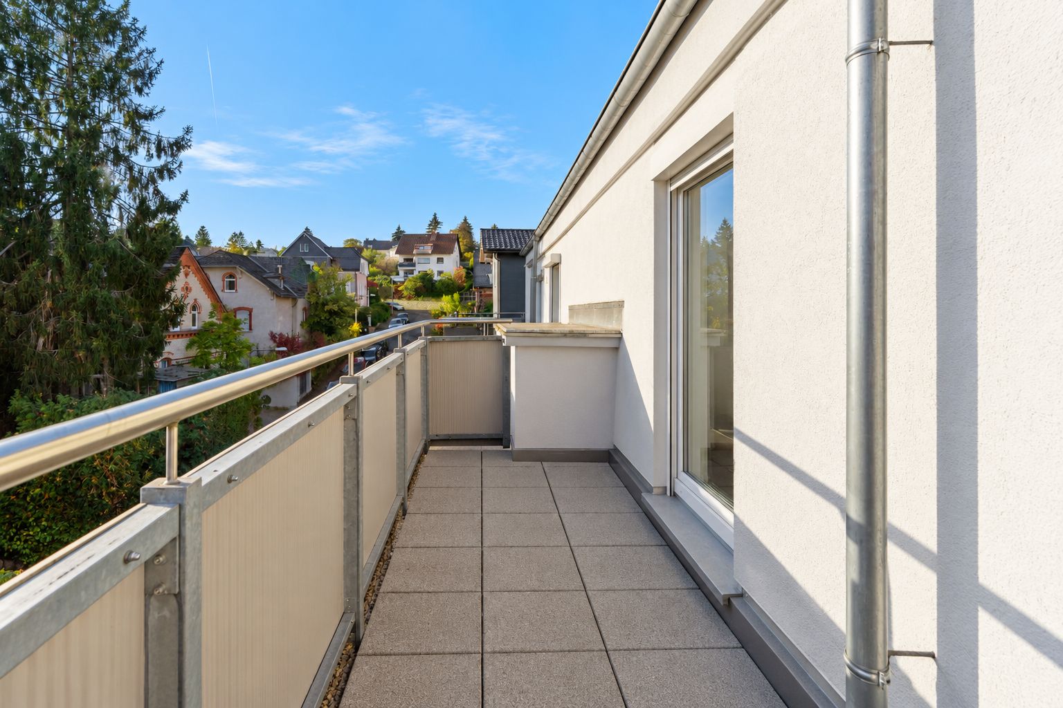 Heller Balkon mit Fliesenboden und Metallgeländer, Blick auf Nachbarhäuser und Bäume.
