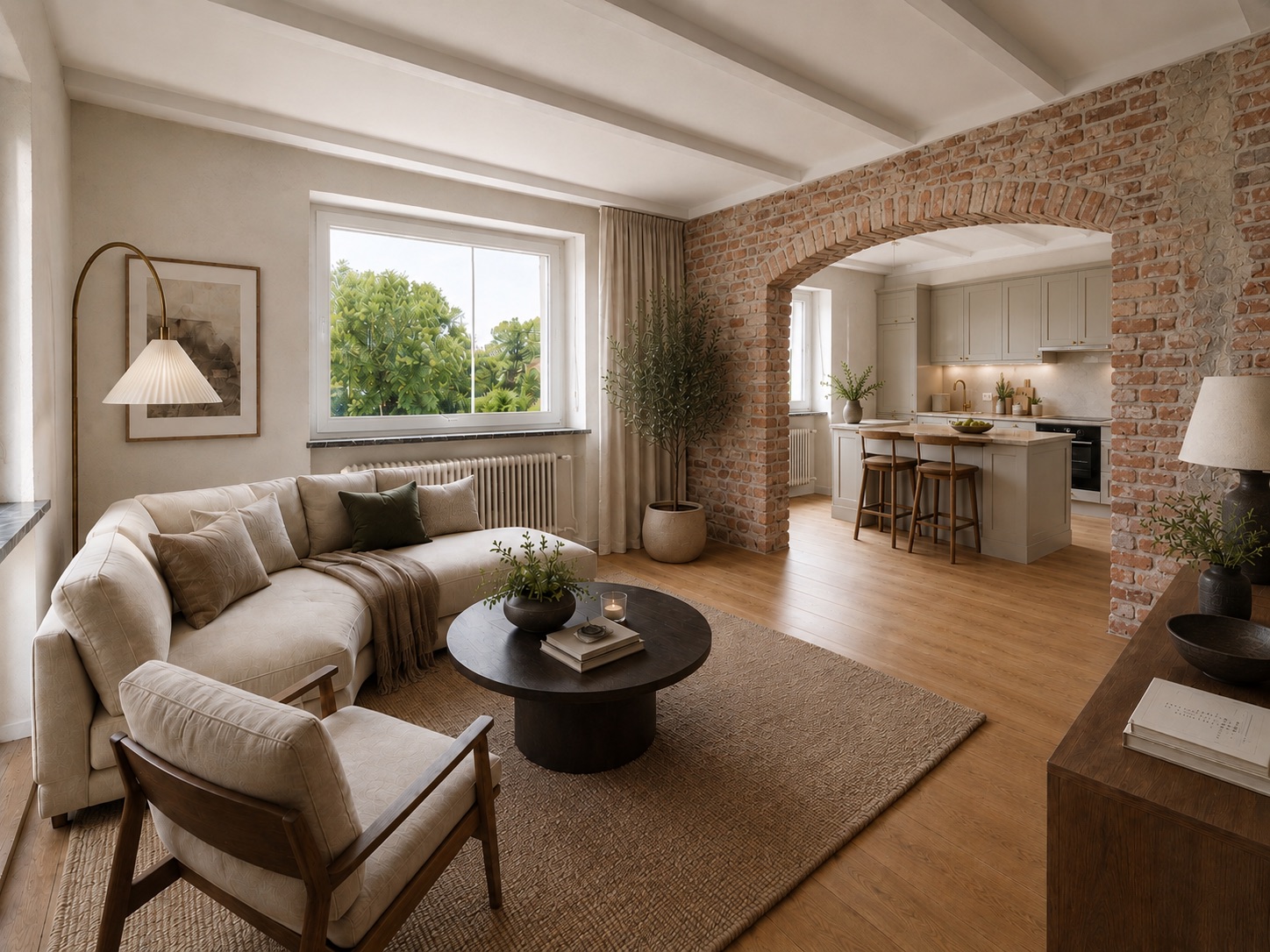 Helles Wohnzimmer mit Sofa, Sessel und rundem Tisch, offene Küche hinter gemauertem Bogen.