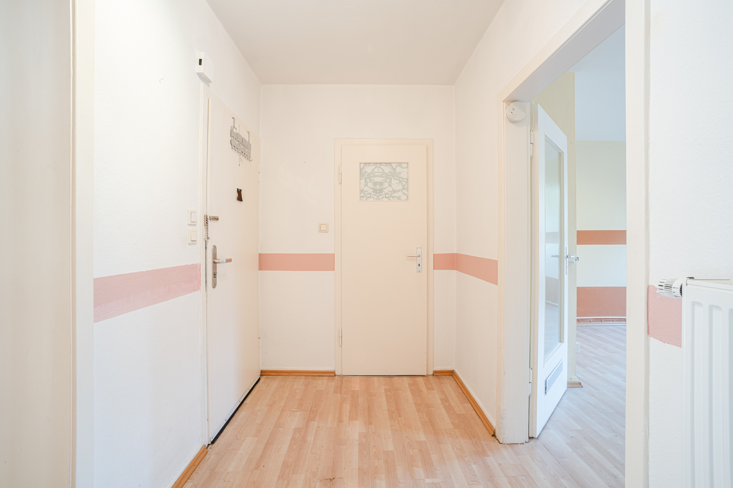 Heller Flur mit Holzboden, weißen Wänden und rosa Streifen, drei Türen sind sichtbar.