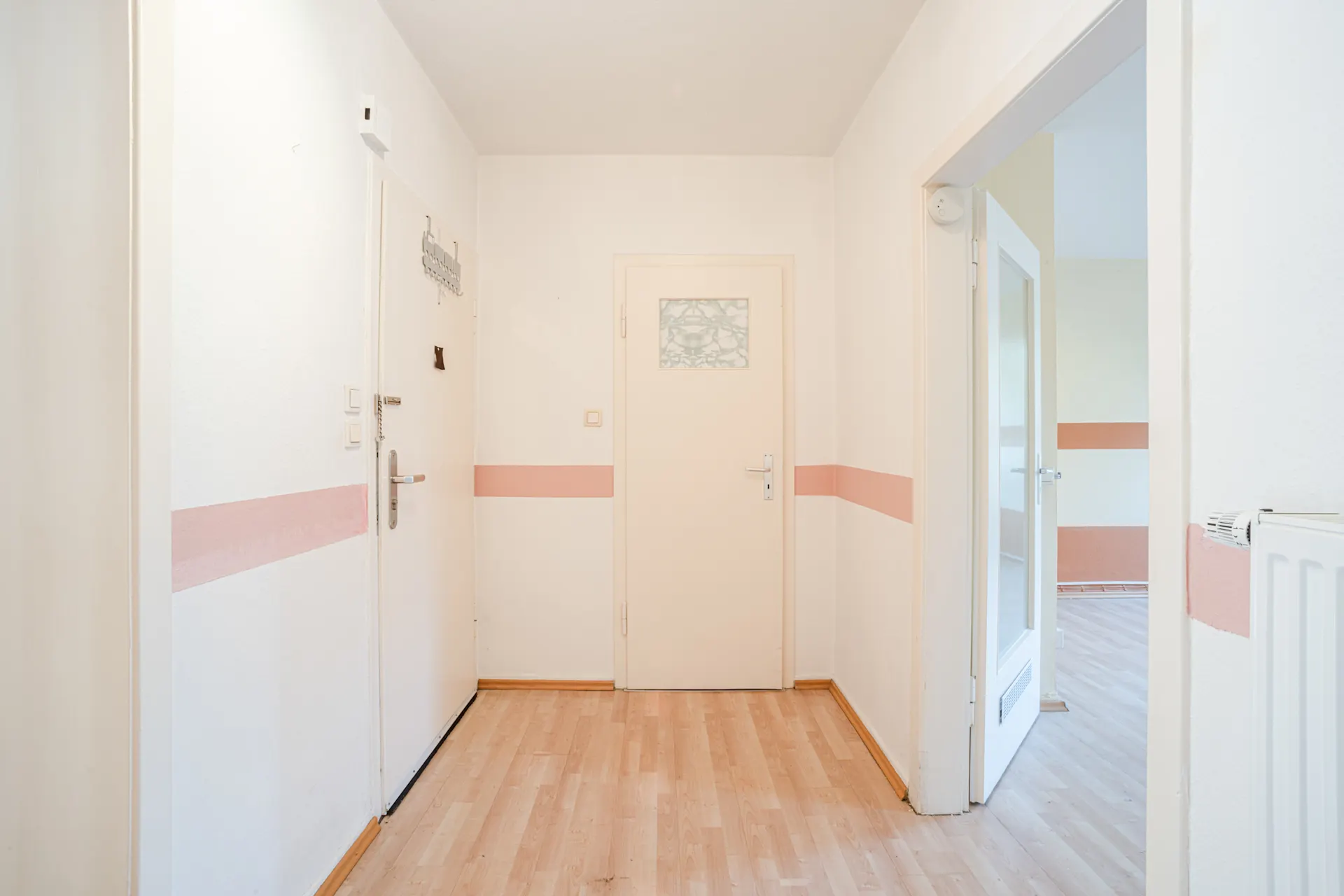 Heller Flur mit Holzboden, weißen Wänden und rosa Streifen, drei Türen sind sichtbar.
