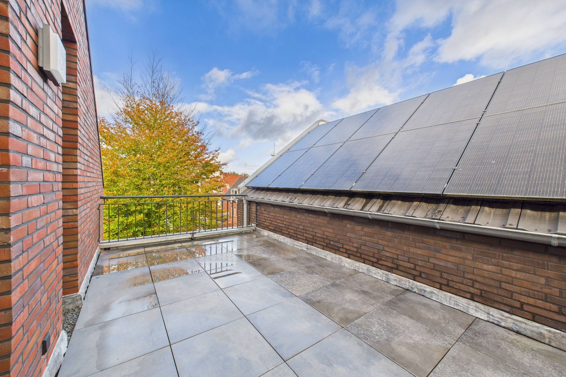 Große Dachterrasse mit grauen Fliesen, Geländer und Solarpanels auf dem Nachbardach.