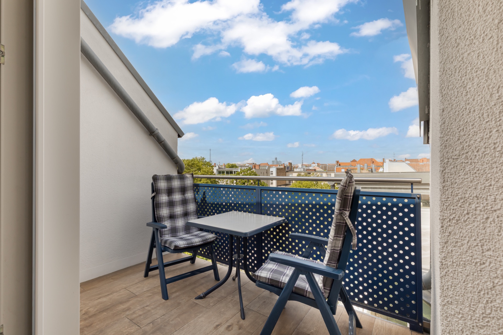 Balkon mit zwei Stühlen und einem kleinen Tisch, Blick auf Dächer und blauen Himmel.