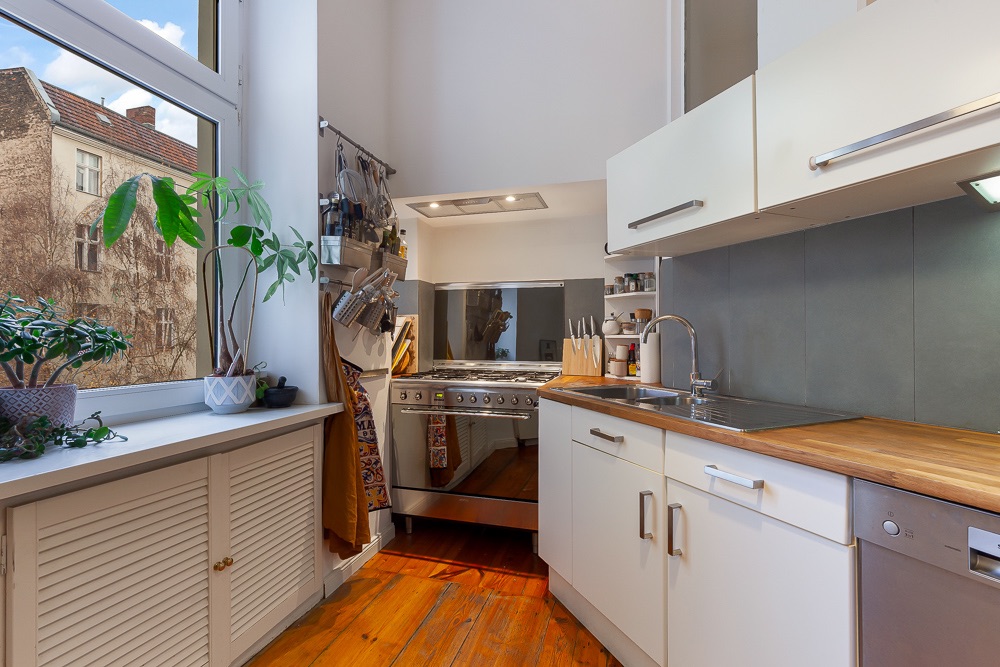Helle Küche mit Holzfußboden, großem Fenster, Pflanzen und modernen weißen Schränken.