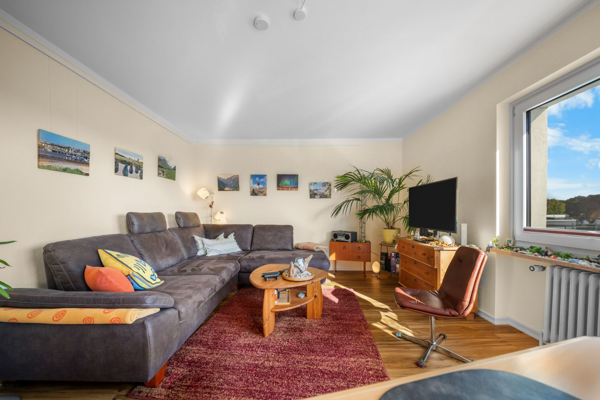 Wohnzimmer mit grauem Ecksofa, Holztisch, Fernseher, großem Fenster und Wandbildern.