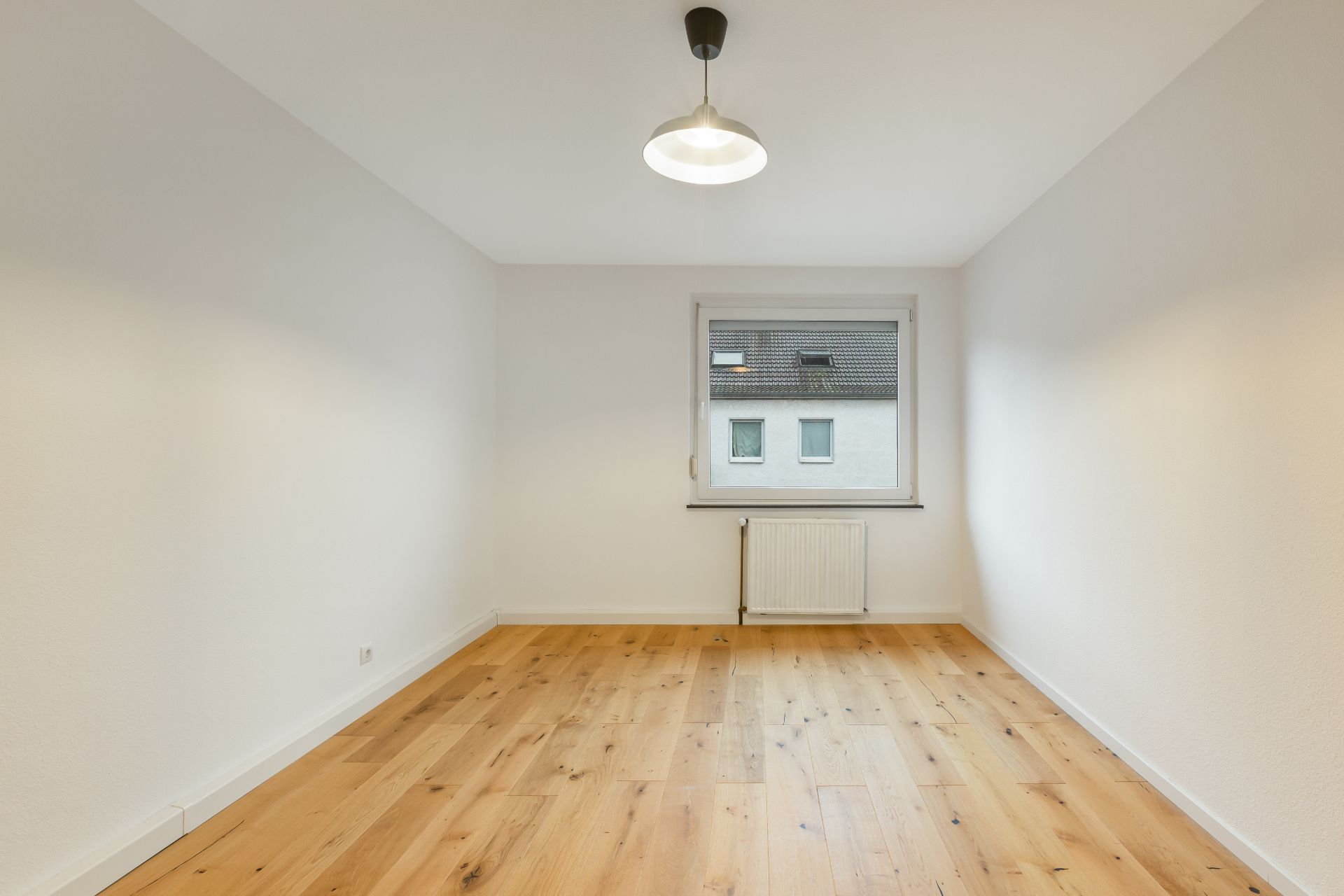 Leerer, heller Raum mit Holzfußboden, weißer Wand, Fenster und Deckenlampe.
