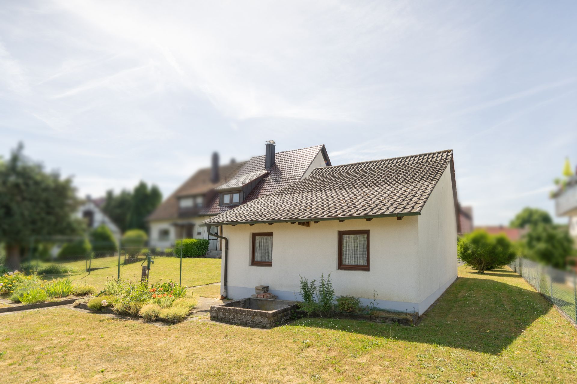 Kleines weißes Haus mit Garten und Blumenbeet auf einer grünen Wiese.