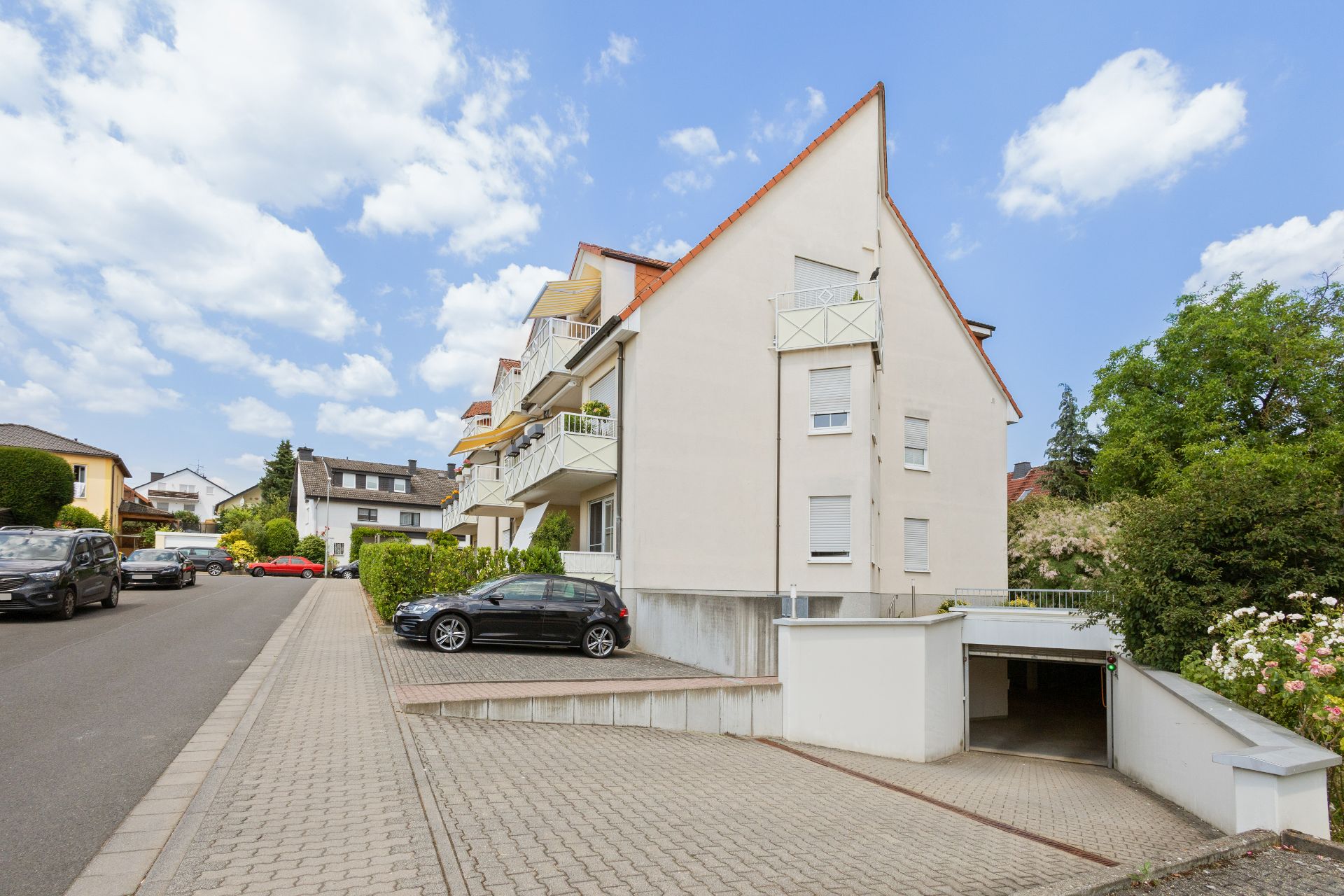 Mehrfamilienhaus mit Balkonen, Garagenzufahrt und geparkten Autos an einer ruhigen Straße.