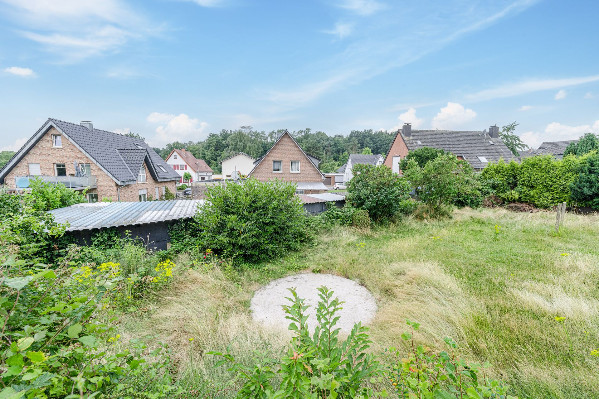 Überwachsener Garten mit runden Sandfläche, Büschen und benachbarten Wohnhäusern im Hintergrund.