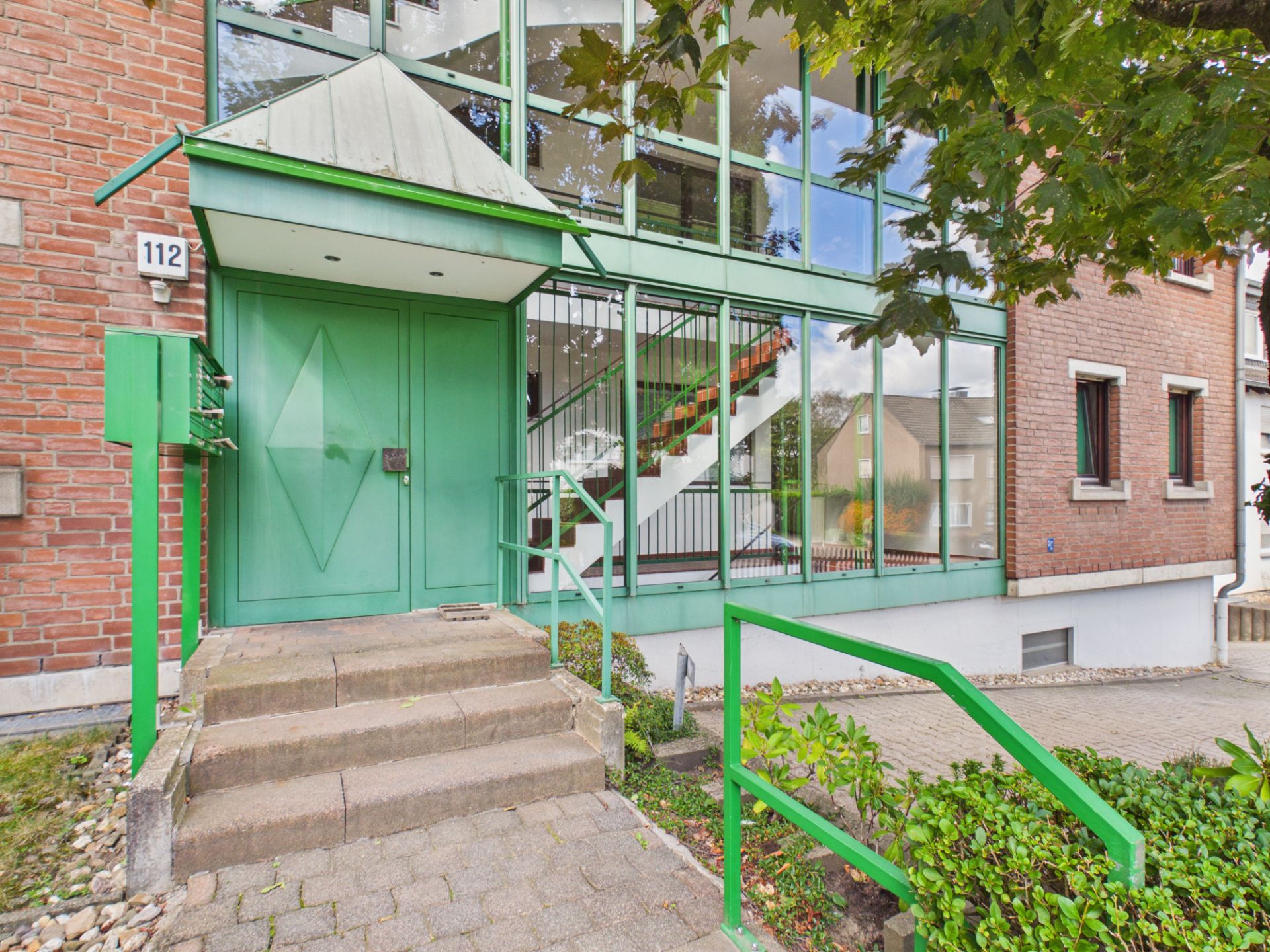 Eingang eines Wohnhauses mit grüner Tür, Treppe und Glasfassade.