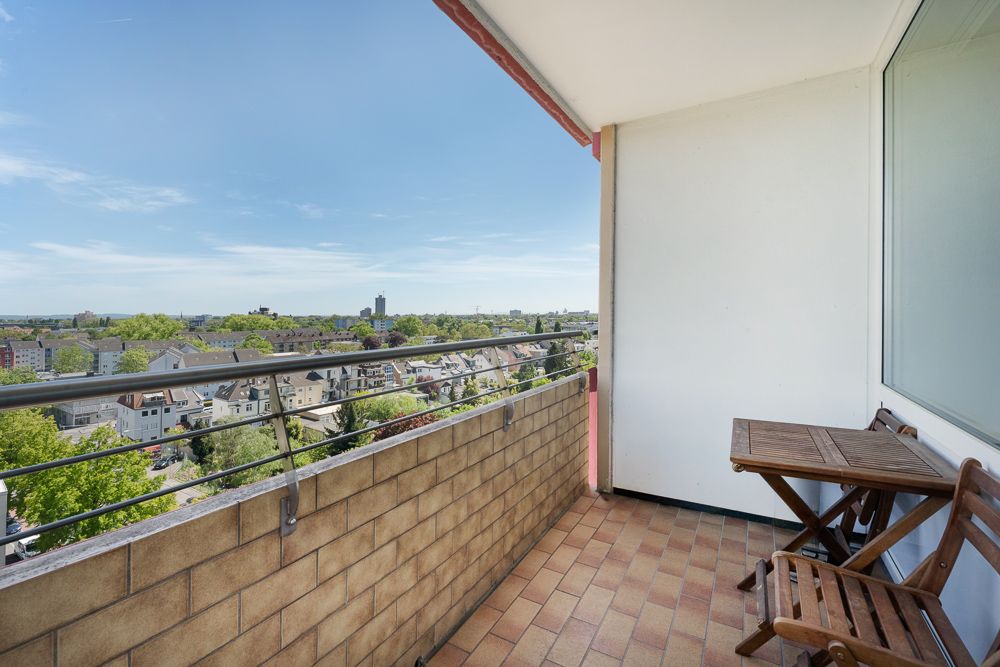 Balkon mit zwei Holzstühlen und Tisch, Blick über Stadt und grüne Bäume.