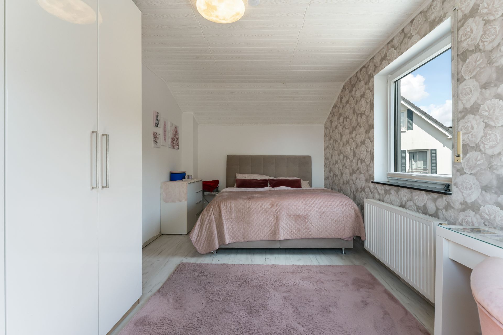 Helles Schlafzimmer mit Doppelbett, Kleiderschrank, Schreibtisch und großem Fenster.
