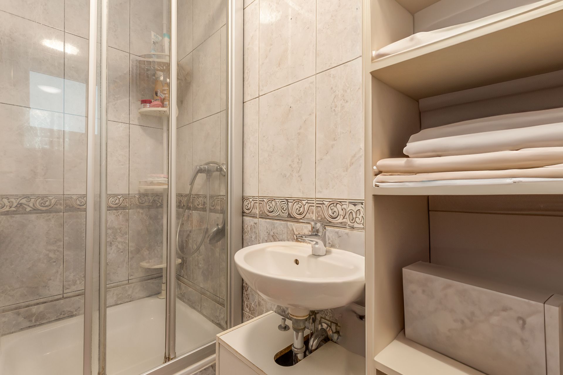 Kleines Badezimmer mit Dusche, Waschbecken und Regal mit gefalteten Handtüchern.