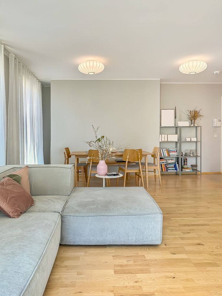 Helles Wohnzimmer mit grauem Sofa, Esstisch, Holzstühlen und Regal auf Holzboden.