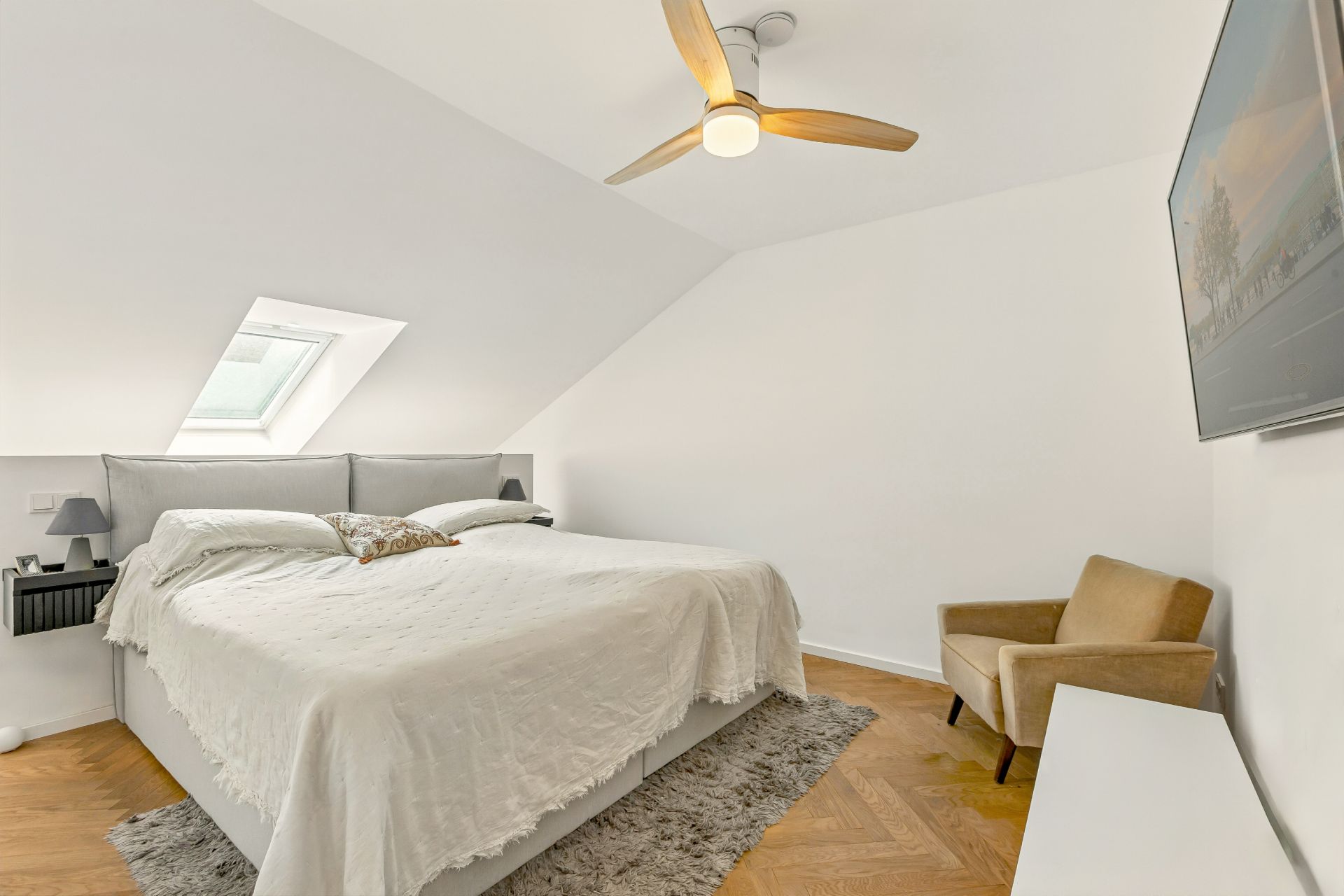 Helles Schlafzimmer mit Doppelbett, Sessel, Teppich, Deckenventilator und Dachfenster.