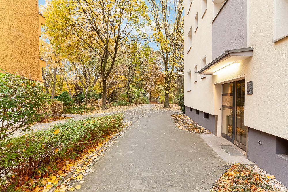 Eingang eines Wohngebäudes mit Fußweg, Bäumen und herbstlichem Laub.