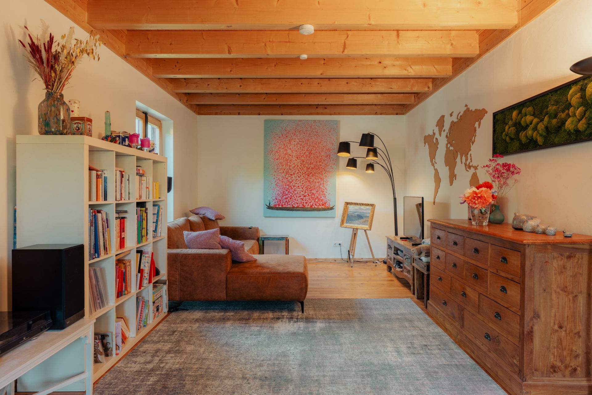 Wohnzimmer mit braunem Sofa, Bücherregal, Teppich und Holzbalkendecke.