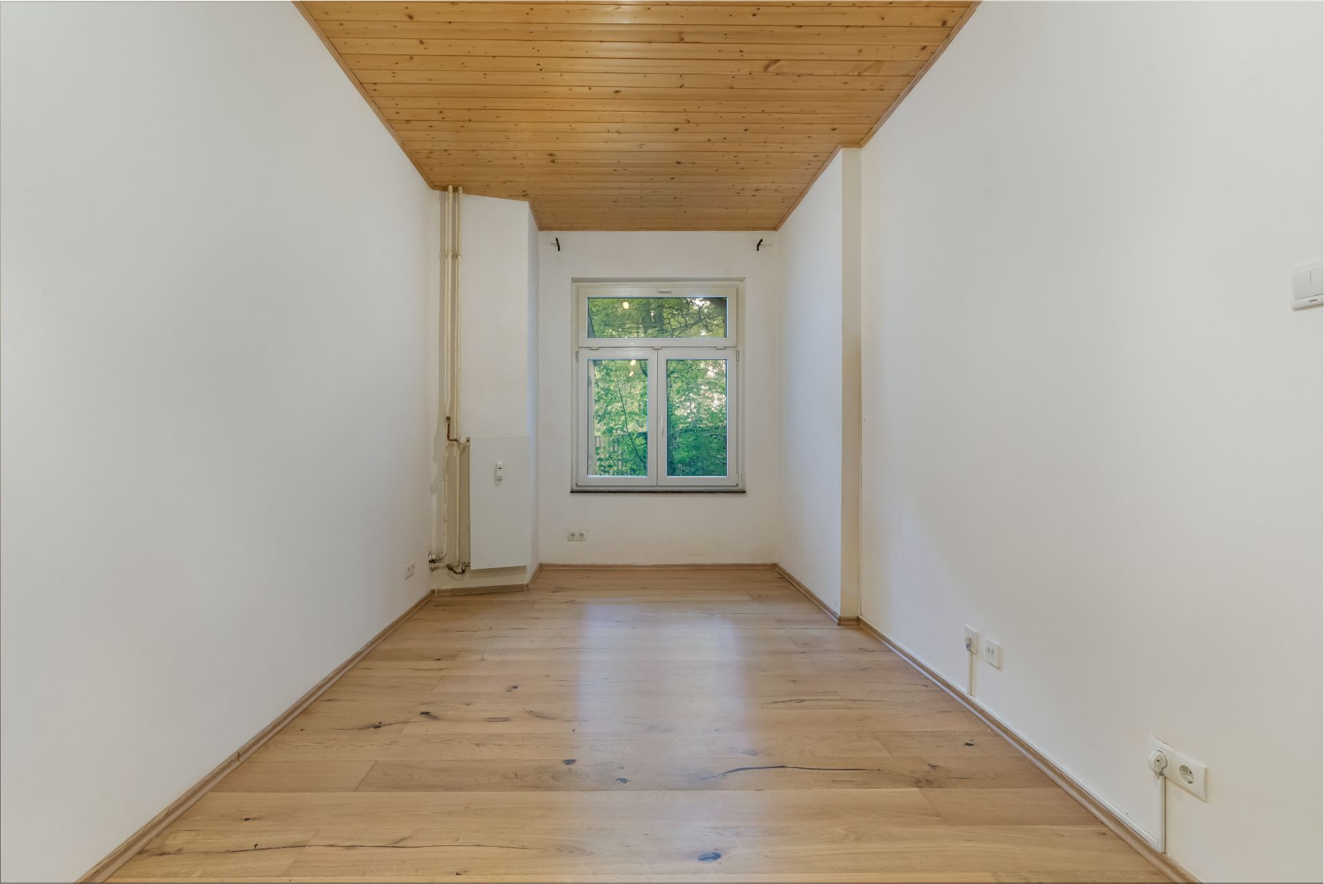 Leerer Raum mit Holzdecke, Holzboden und einem Fenster mit Blick ins Grüne.