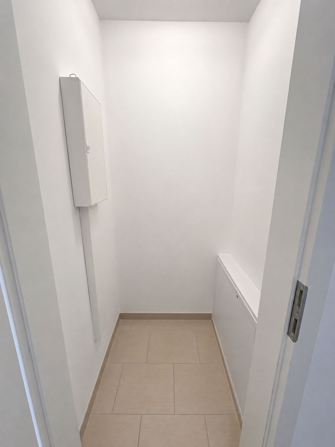 Kleiner, leerer Raum mit Fliesenboden und weißen Wänden, an der Wand ist ein Kasten montiert.