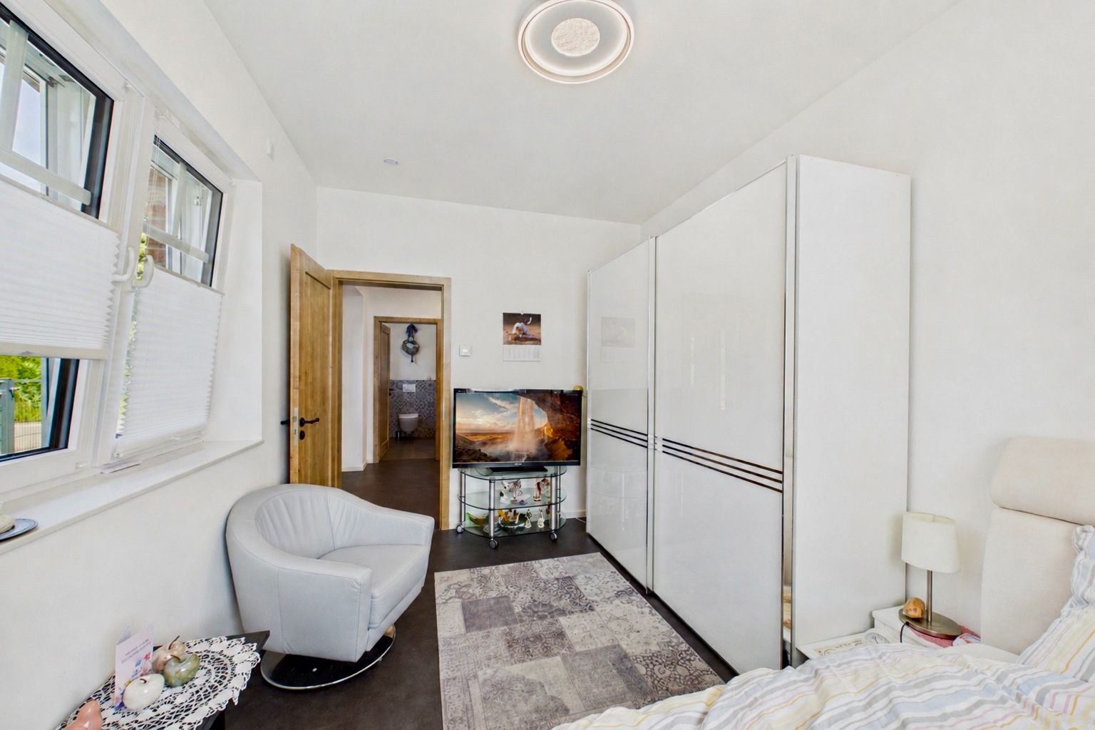 Helles Schlafzimmer mit Teppich, Kleiderschrank, Sessel, Fernseher und Fenster.