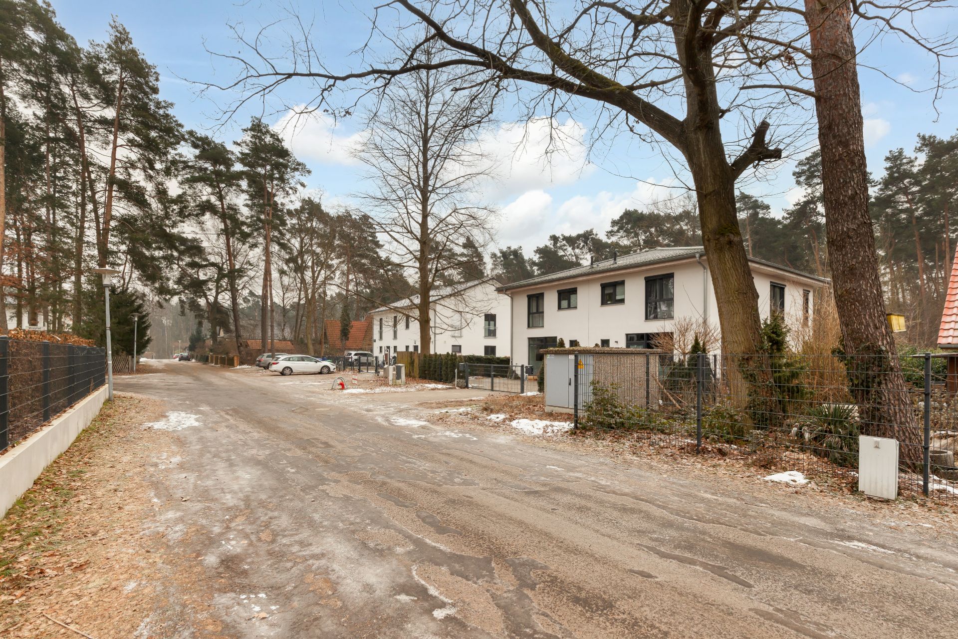 Wohnstraße mit modernen weißen Häusern, Bäumen und etwas Schnee am Straßenrand.