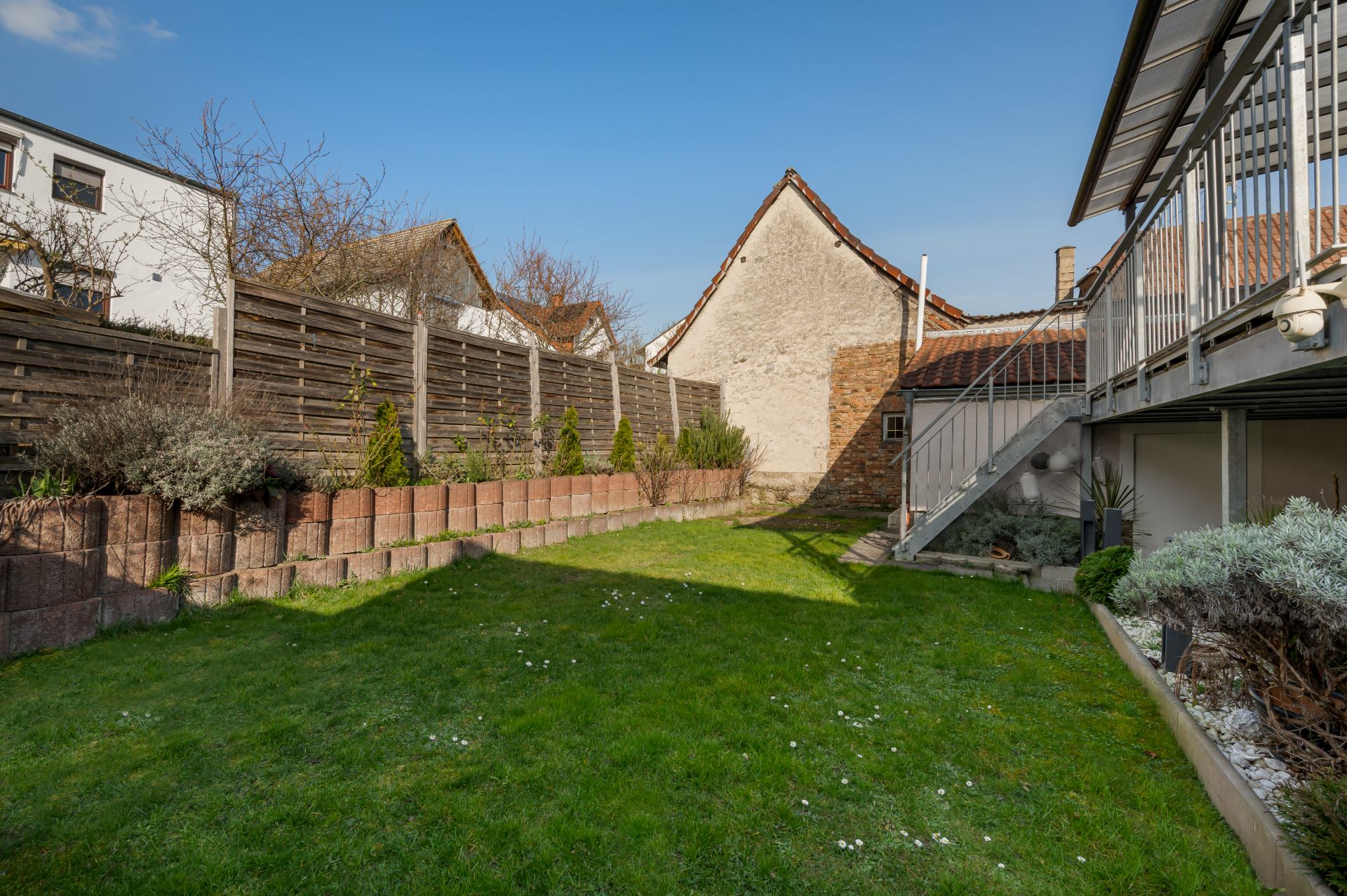 Grüner Garten mit Rasenfläche, Holzlattenzaun und Terrasse mit Treppe an einem Wohnhaus.