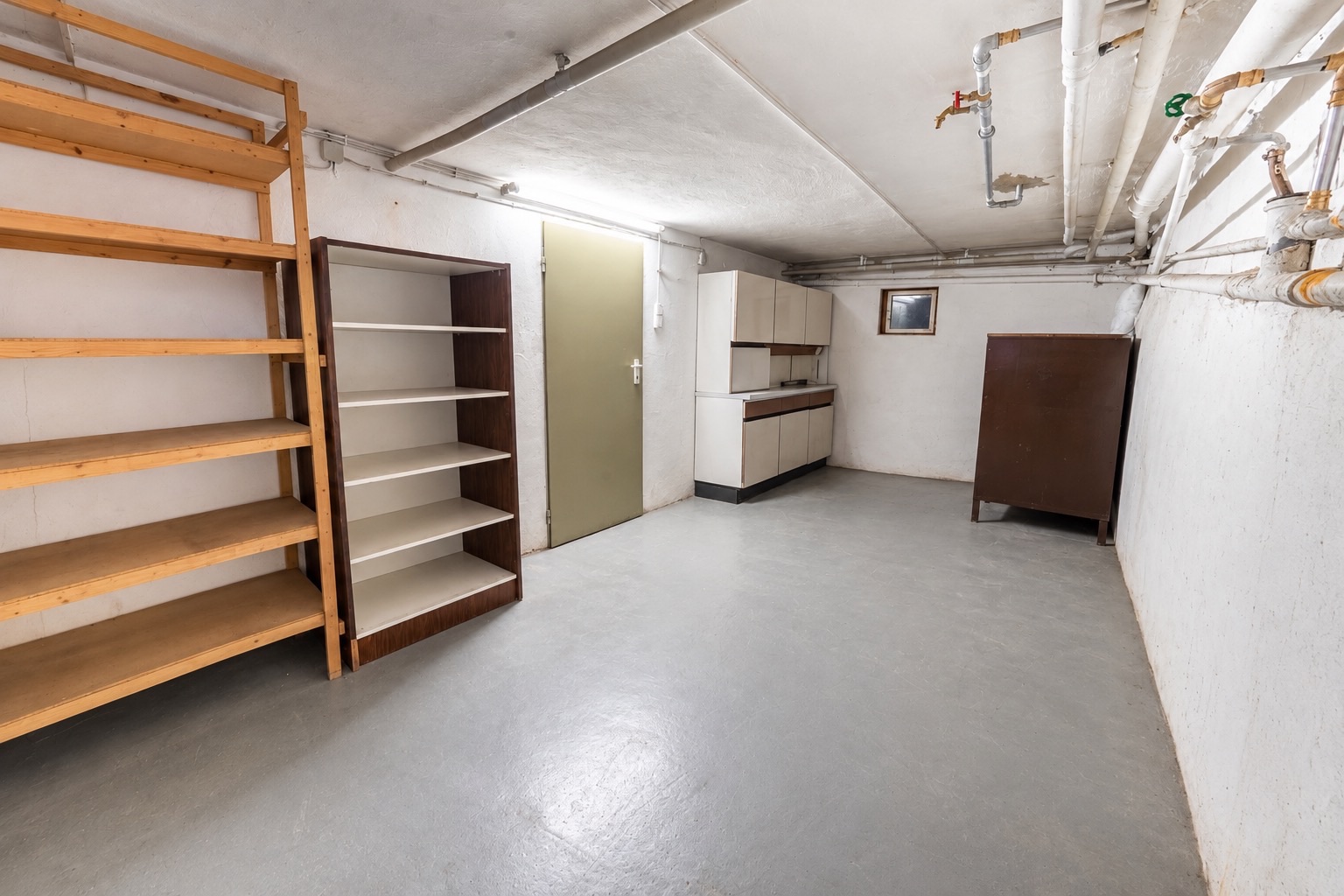 Kellerraum mit Holzregalen, Schrank, Kommode und sichtbaren Rohrleitungen an der Decke.