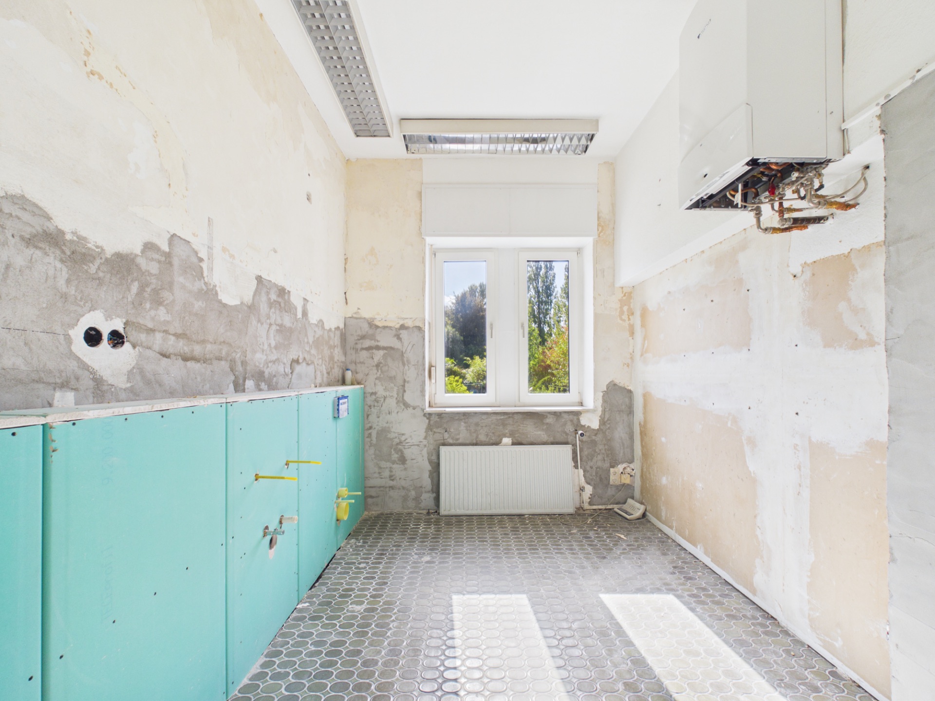 Leerer, teils renovierter Raum mit Fenster, Heizkörper und sichtbaren Rohren.