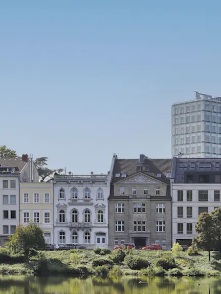 Mehrere Wohngebäude stehen nebeneinander an einem See, im Vordergrund sind Bäume und Wasser.