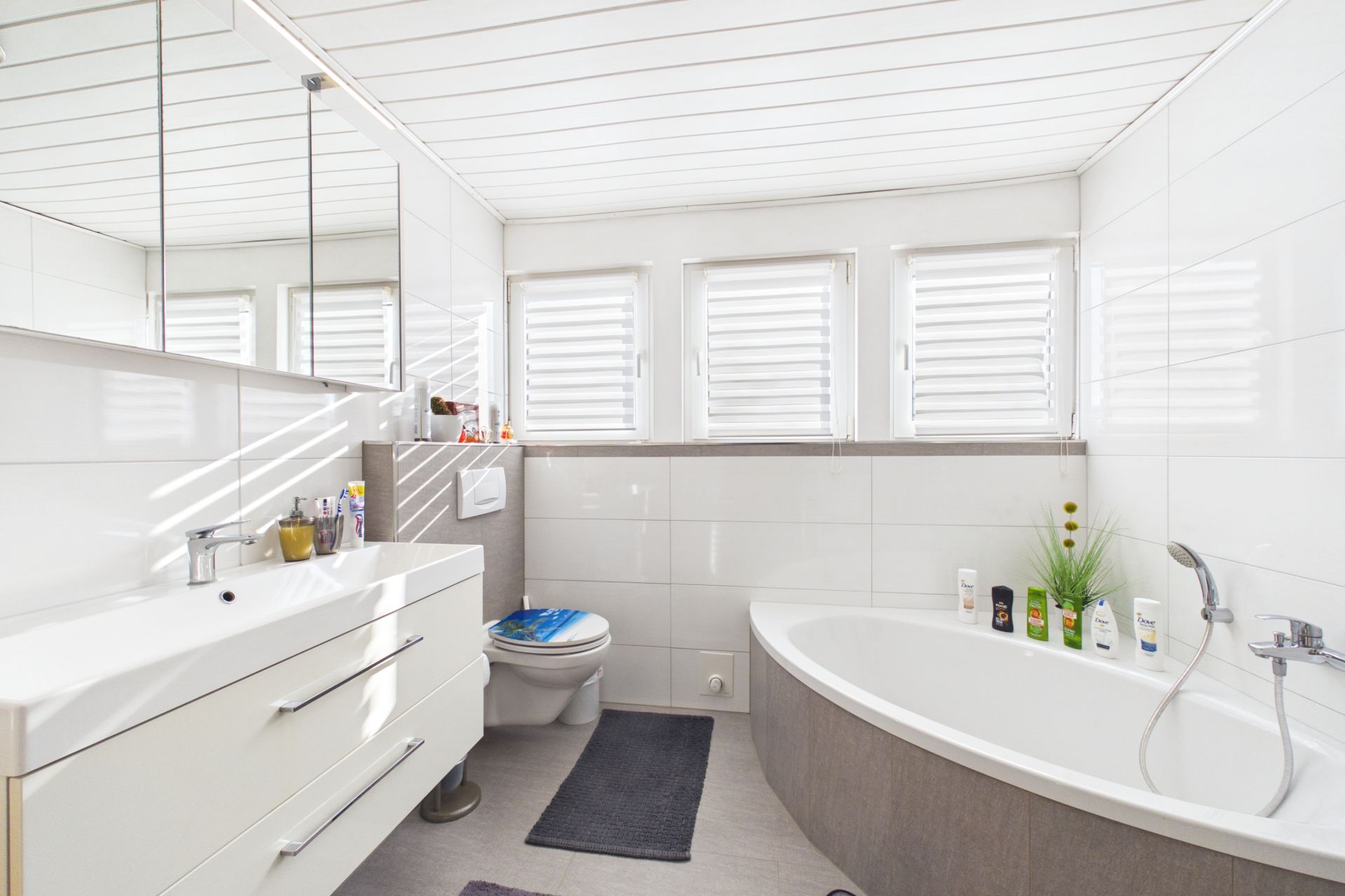 Helles Badezimmer mit Badewanne, Waschbecken, Toilette und Fenstern mit Jalousien.