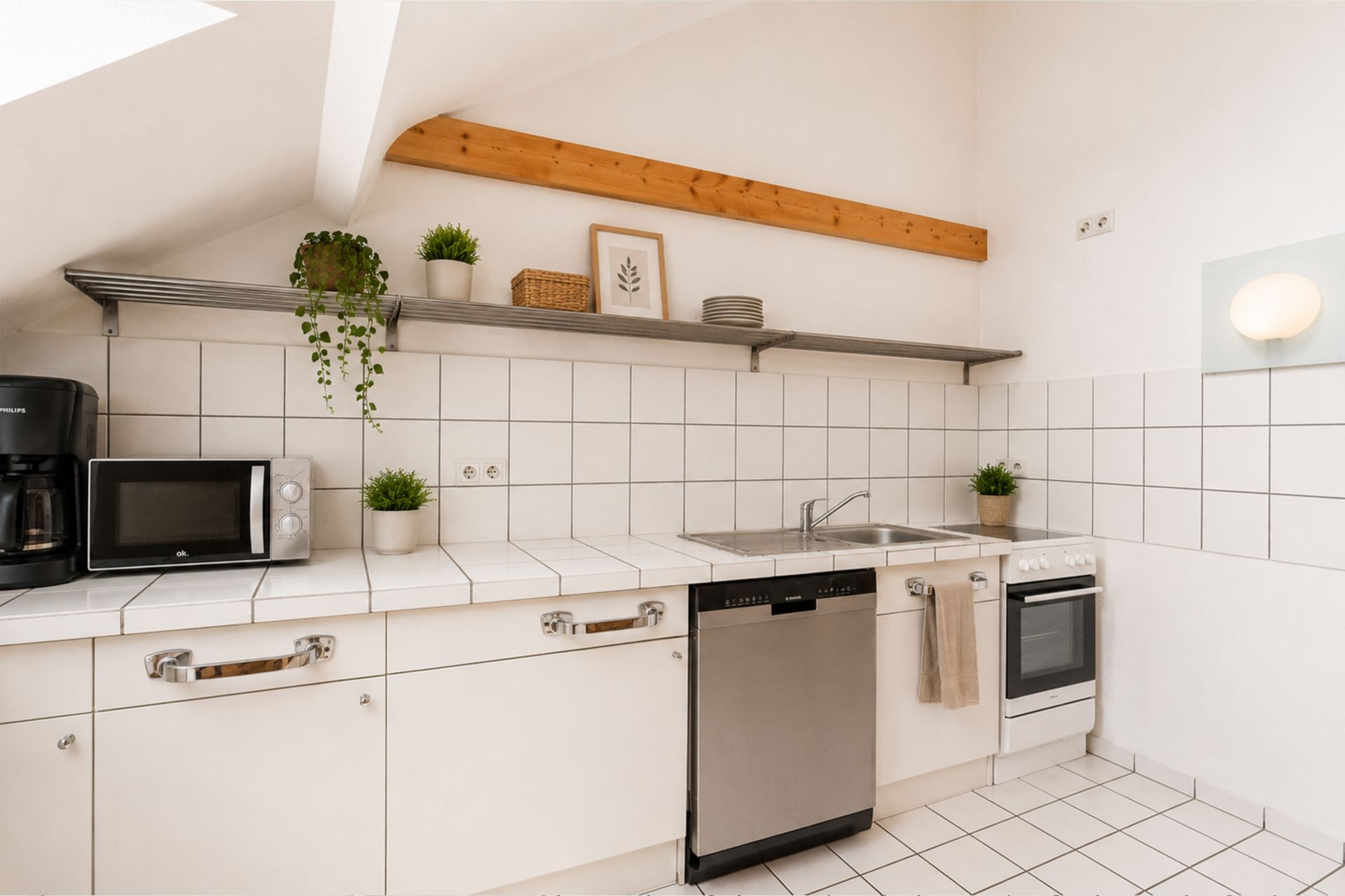 Helle Küche mit weißen Fliesen, Spüle, Backofen, Geschirrspüler und Regal mit Pflanzen und Geschirr.