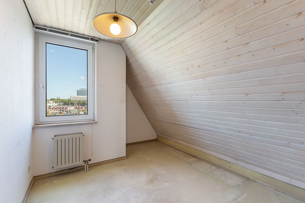 Leeres Zimmer mit schräger Holzdecke, Fenster und Heizung an der Wand.