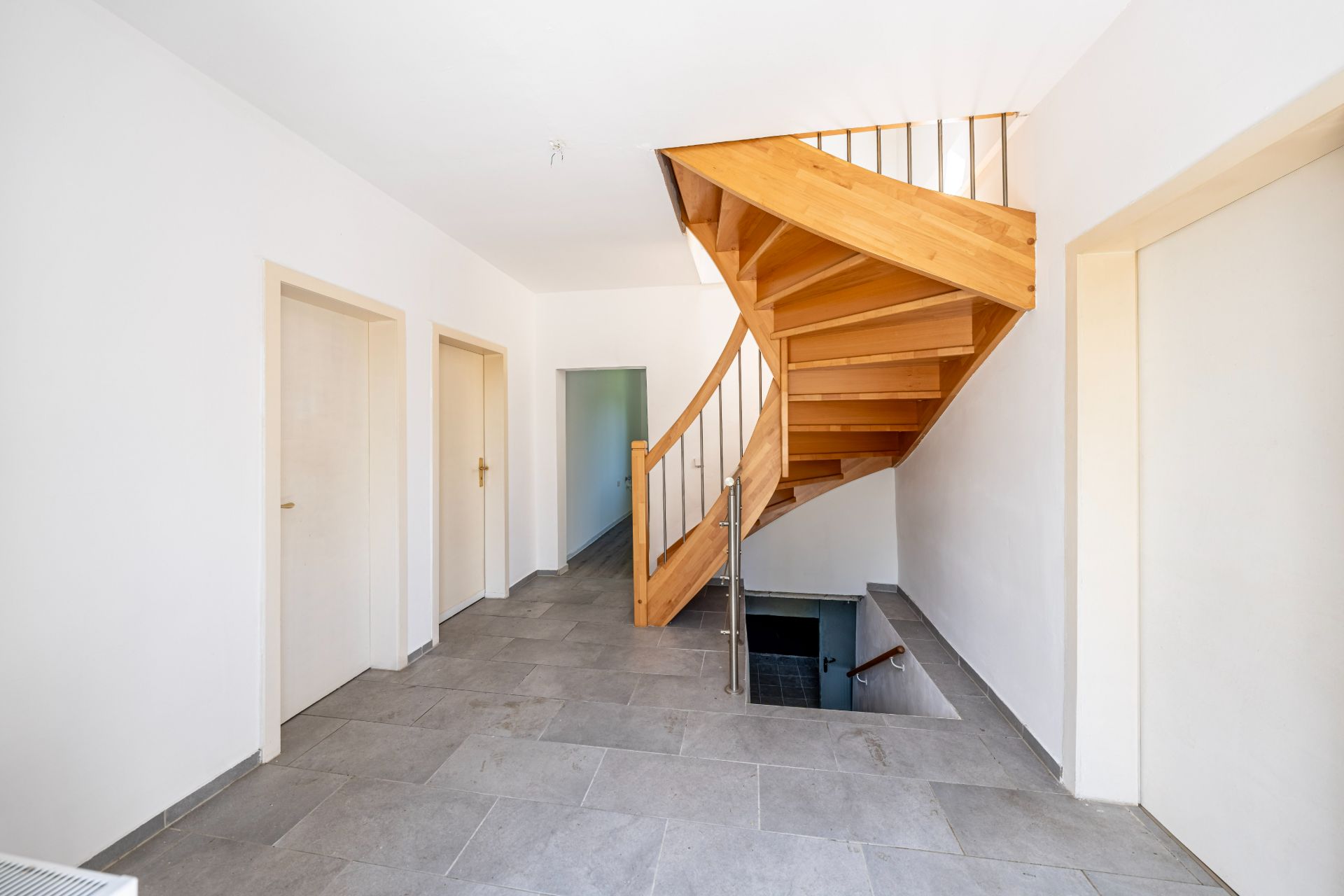 Flur mit grauem Fliesenboden, vier Türen und einer Holztreppe nach oben und unten.
