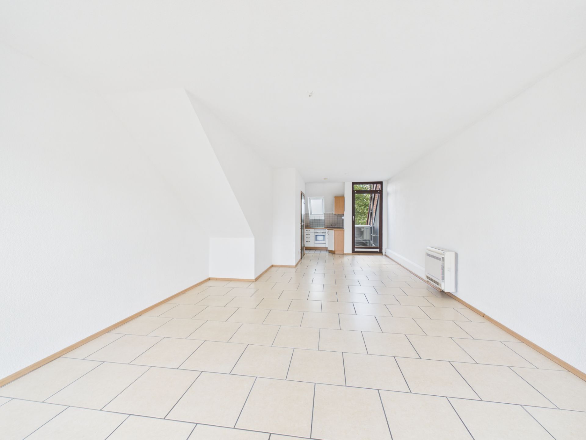 Heller, leerer Wohnraum mit Fliesenboden, offener Küche und Zugang zu einem Balkon.