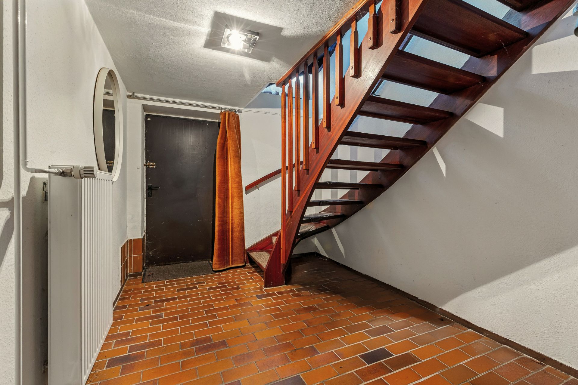 Flur mit braunen Fliesen, Holztreppe, Tür und rundem Spiegel an der Wand.