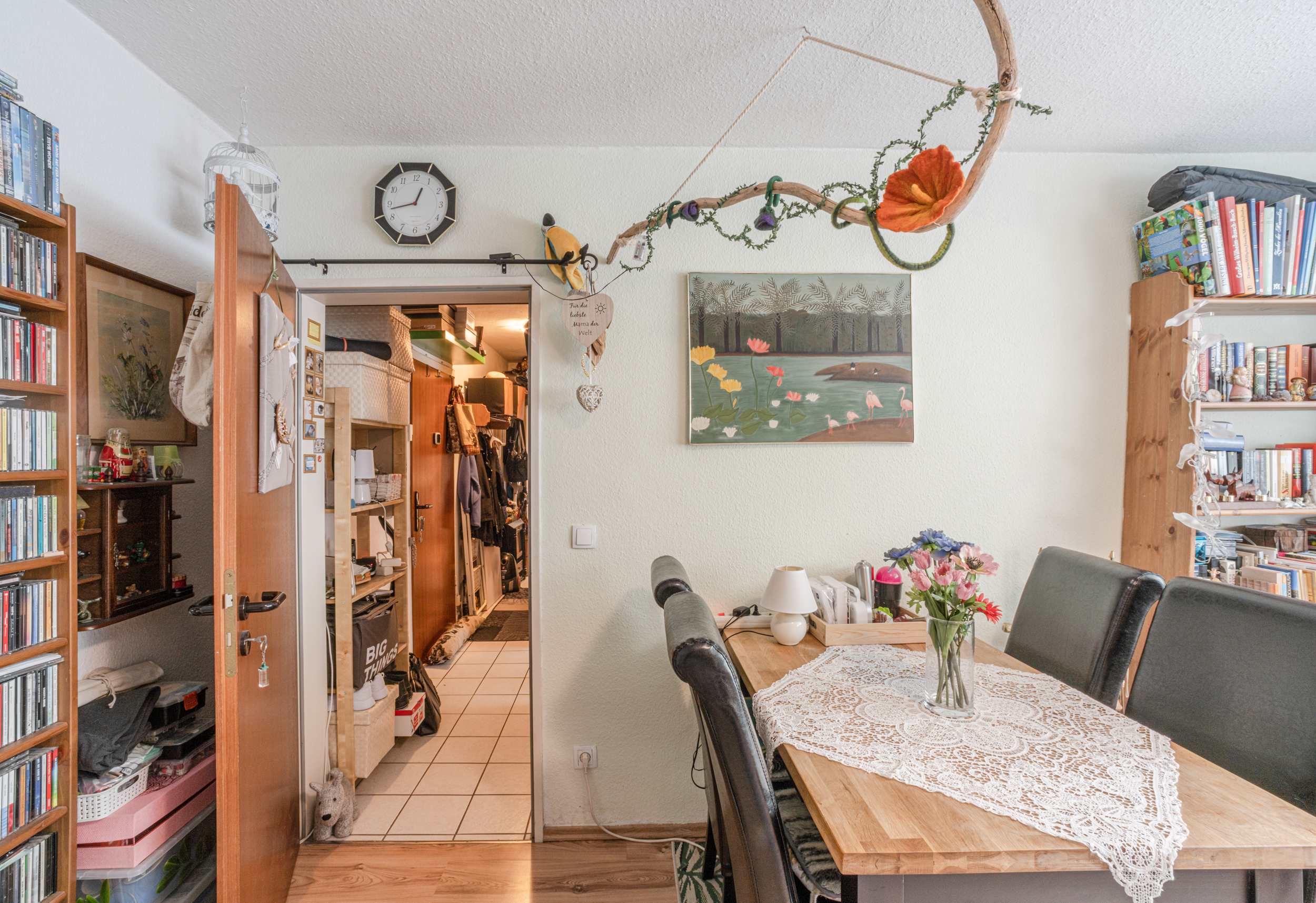 Essbereich mit Holztisch, Blumenstrauß, Bücherregal und Blick in einen Flur mit offenen Regalen.