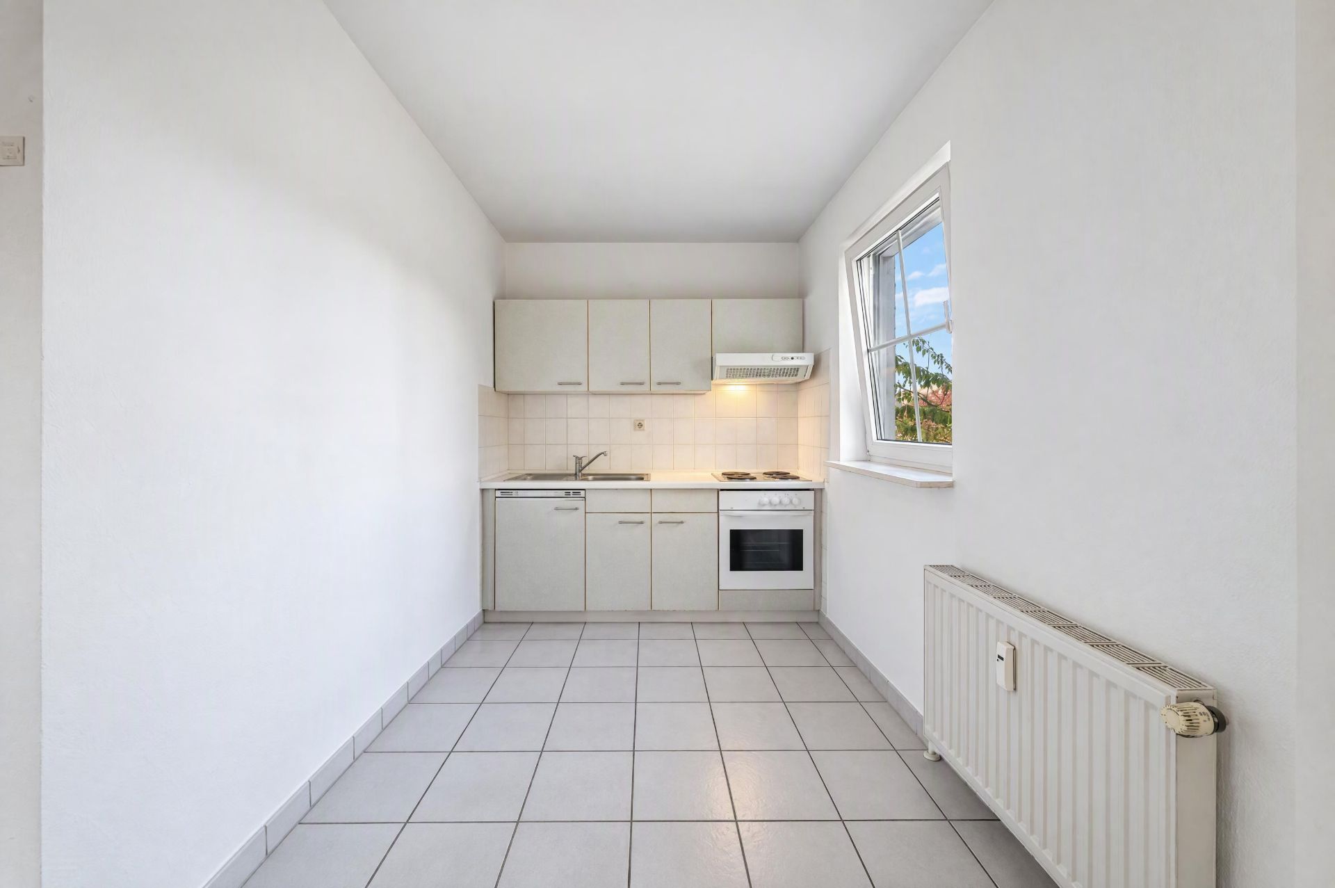 Helle Einbauküche mit weißen Schränken, Fenster und Heizkörper, gefliester Boden.