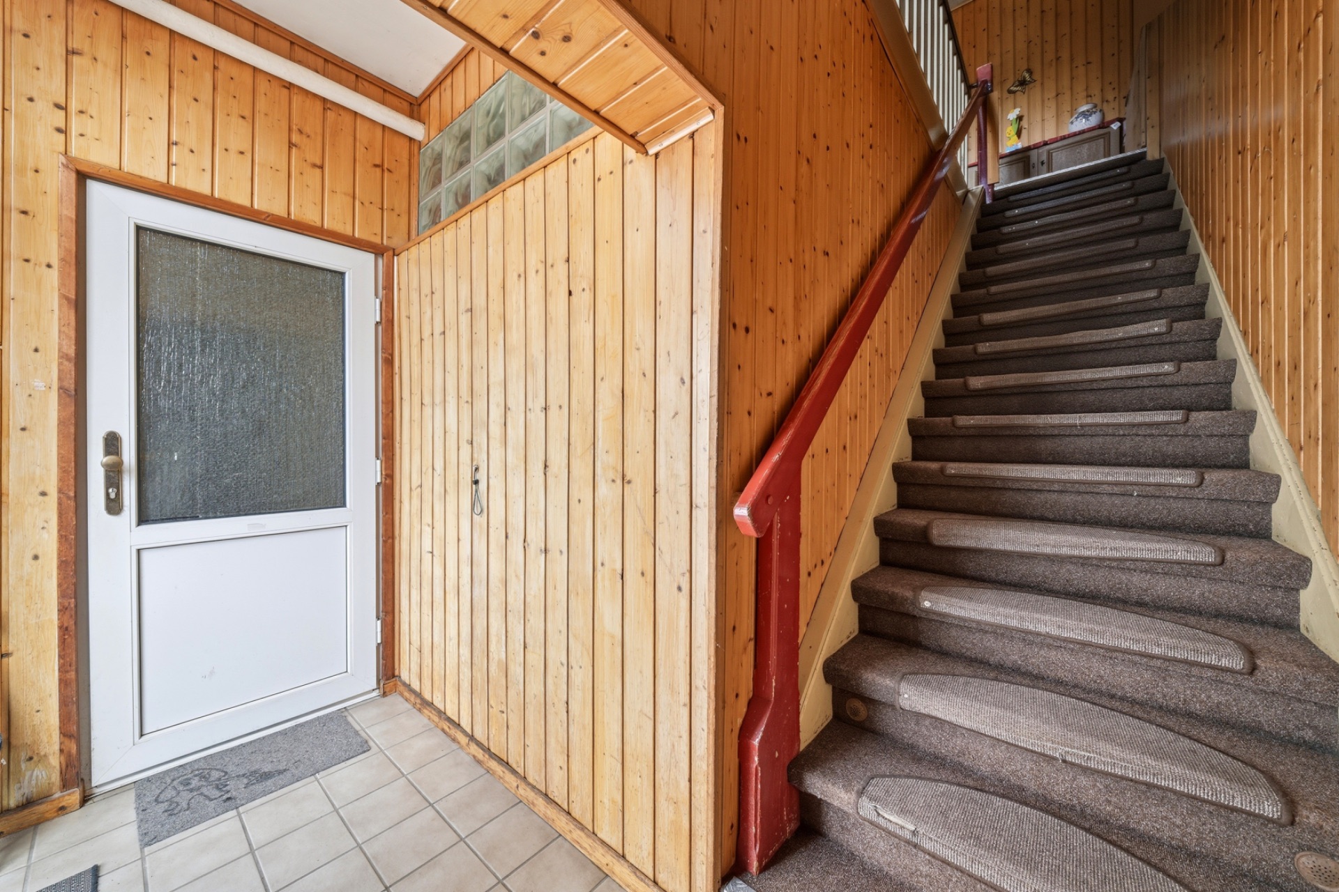 Flur mit Holzwänden, weißer Eingangstür und Teppichtreppe mit rotem Geländer.