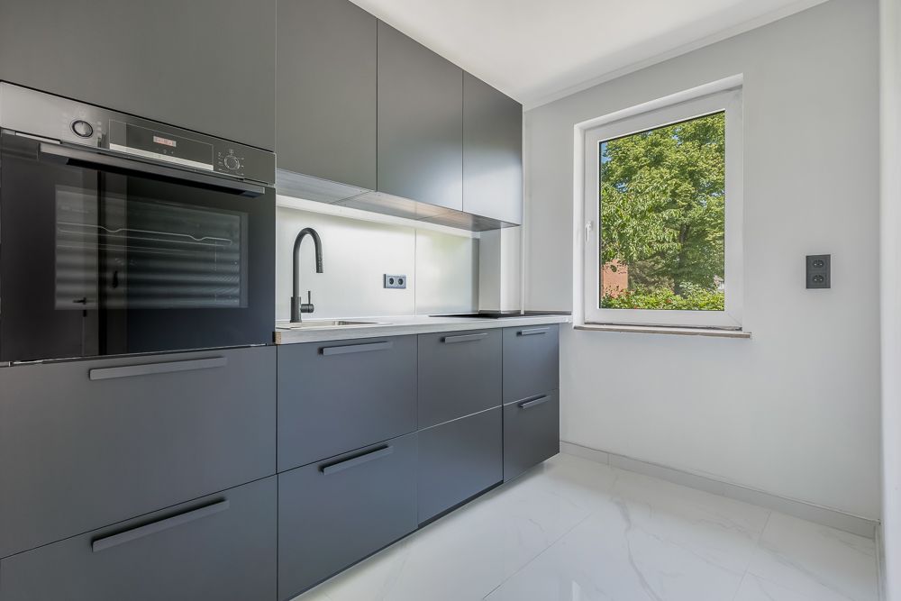 Moderne Küche mit grauen Schränken, Einbauofen, Spüle und Fenster mit Blick ins Grüne.