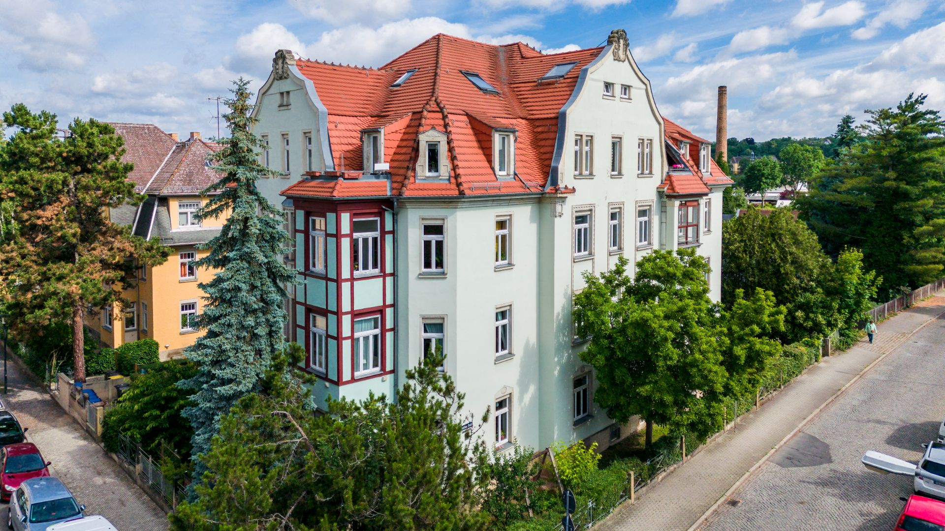 Großes Wohnhaus mit rotem Dach und weißen Fassaden, umgeben von Bäumen an einer ruhigen Straße.