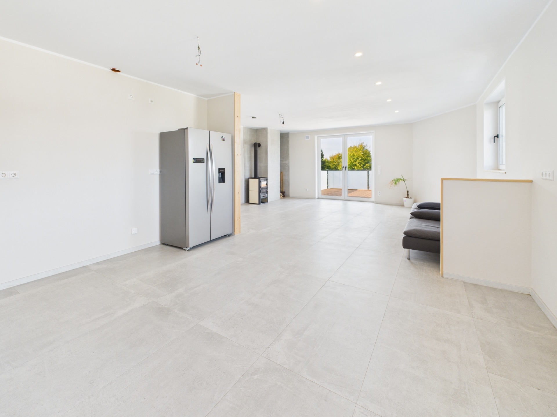 Heller, leerer Wohnraum mit Kühlschrank, kleiner Sitzecke und großen Fenstern zur Terrasse.