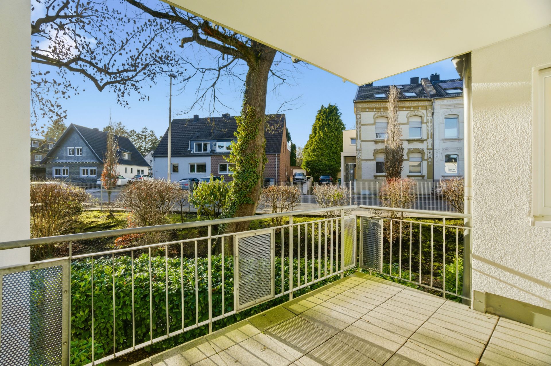 Blick von einem Balkon mit Metallgeländer auf eine ruhige Wohnstraße mit Bäumen und Häusern.
