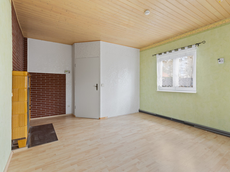 Helles, leeres Zimmer mit Holzdecke, grün gestrichenen Wänden, Fenster und gelbem Kachelofen.