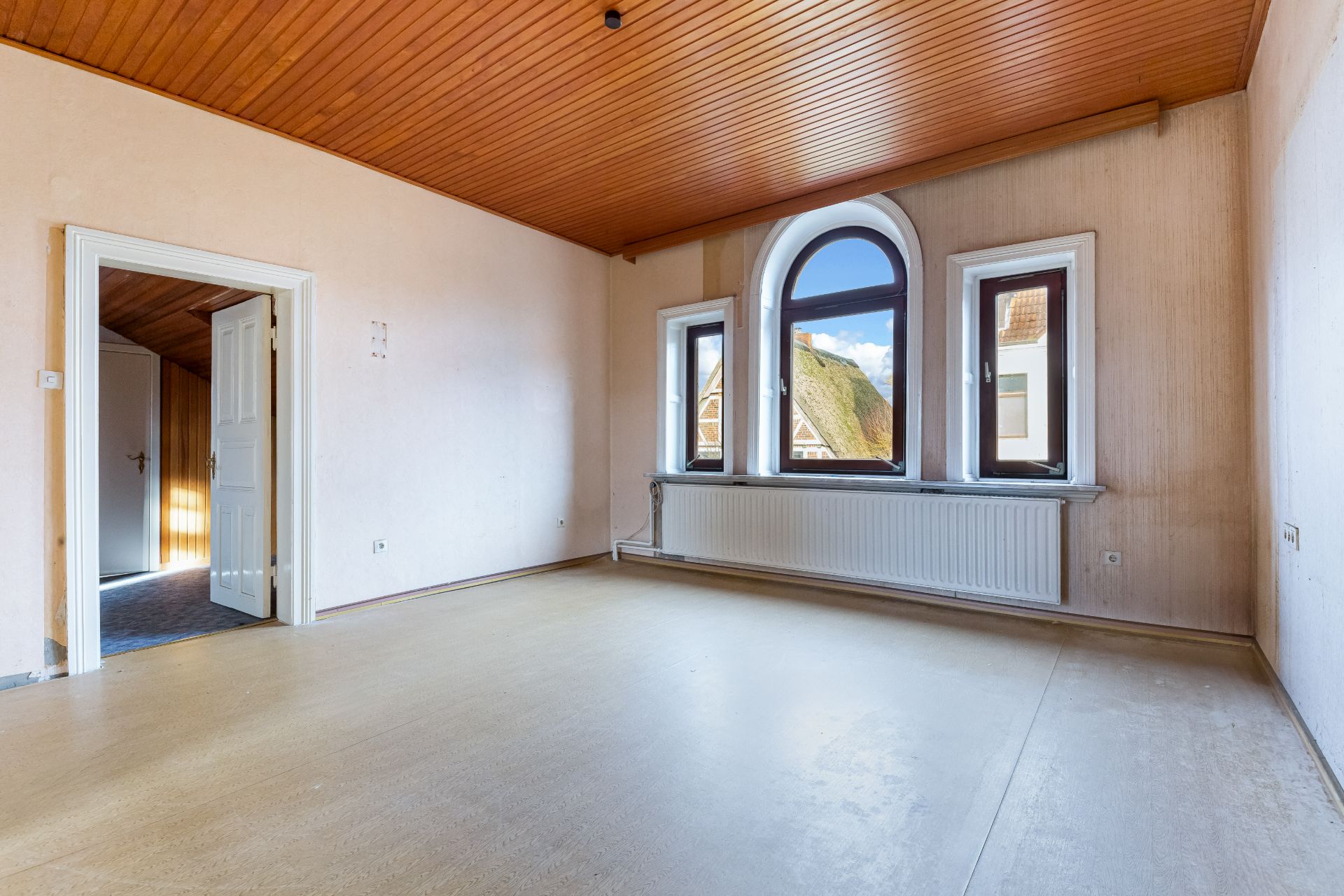 Leeres Zimmer mit Holzdecke, drei Fenstern und offenem Durchgang zu einem weiteren Raum.