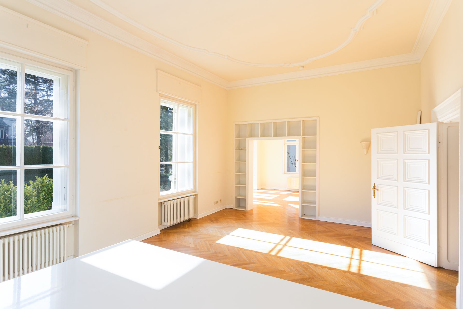 Helles Zimmer mit Parkettboden, großen Fenstern und offener Tür zu einem weiteren Raum.