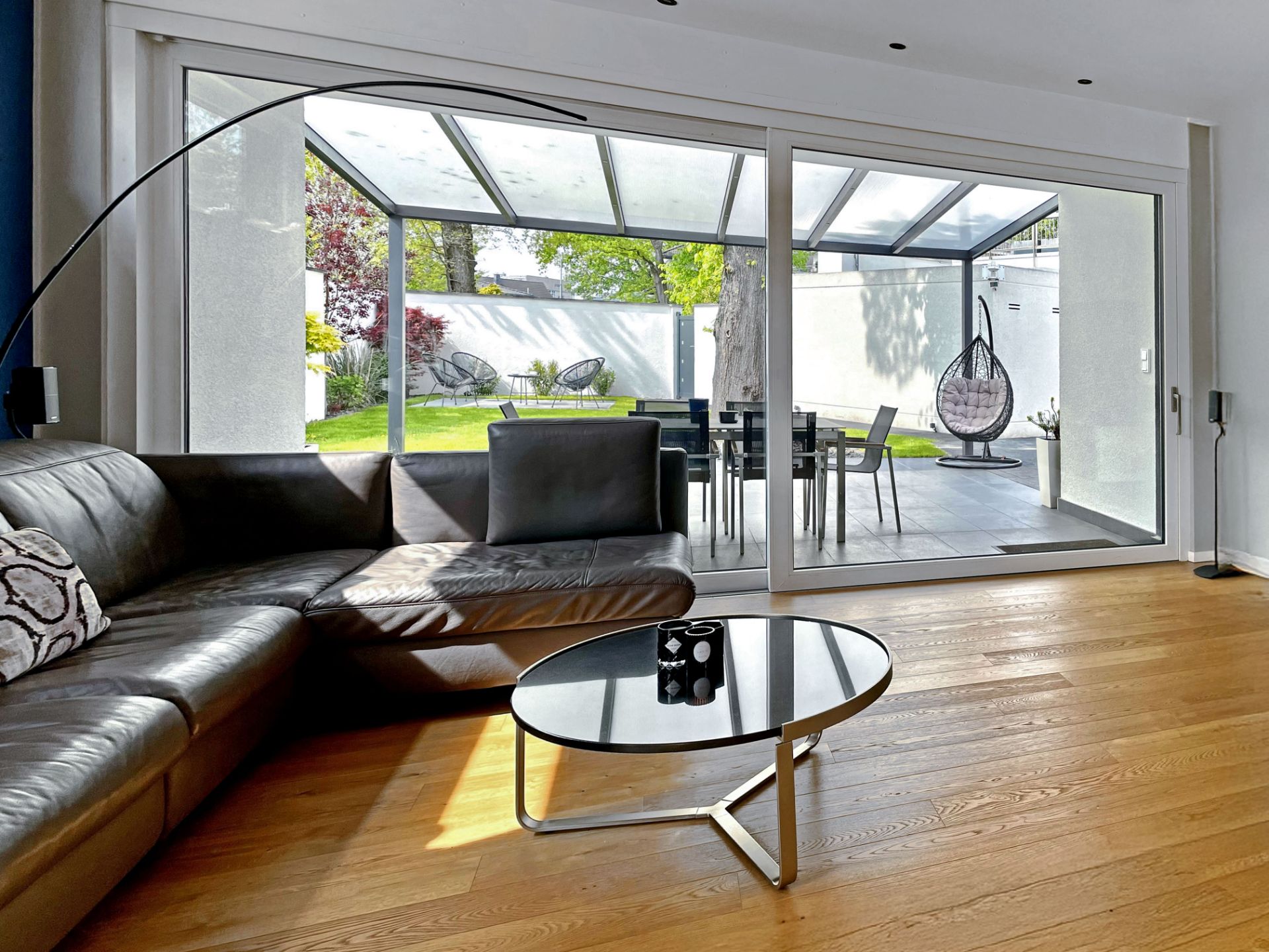 Modernes Wohnzimmer mit Ledersofa, Glastisch und Blick auf Terrasse und Garten durch große Glasschiebetüren.