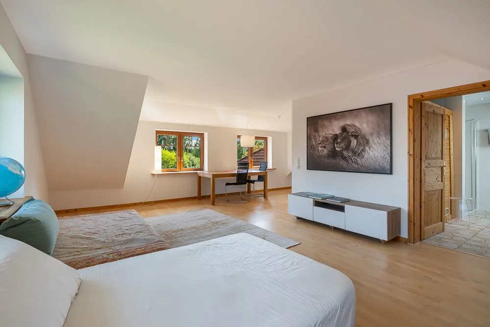 Helles Schlafzimmer mit Holzboden, Bett, Schreibtisch am Fenster und Kommode mit großem Bild an der Wand.