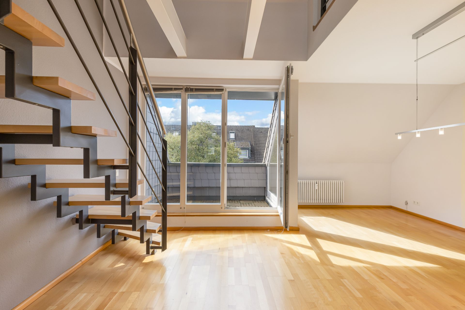 Helles Wohnzimmer mit Holztreppe, großen Fenstern und Zugang zu einem Balkon.