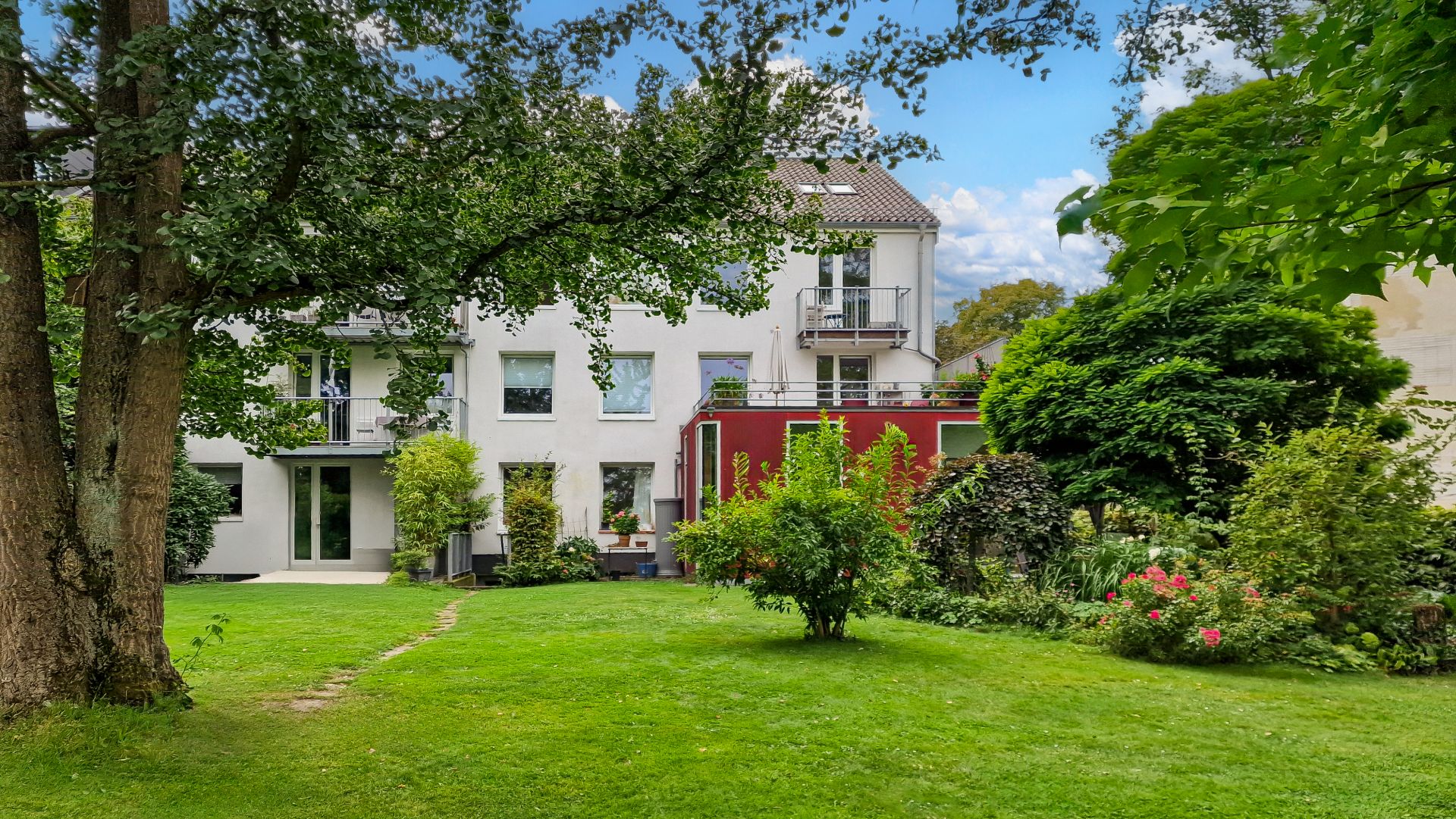 Hinteransicht eines Wohnhauses mit großem Garten, Rasen, Bäumen und blühenden Sträuchern.